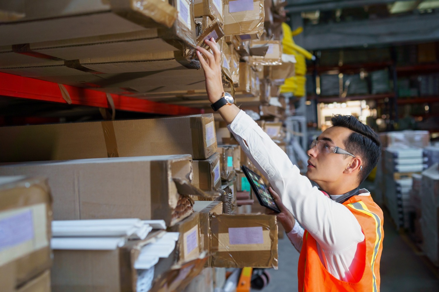 Alt text: Warehouse worker in a high-visibility vest scanning and retrieving boxed inventory from storage shelves in a fulfilment centre.