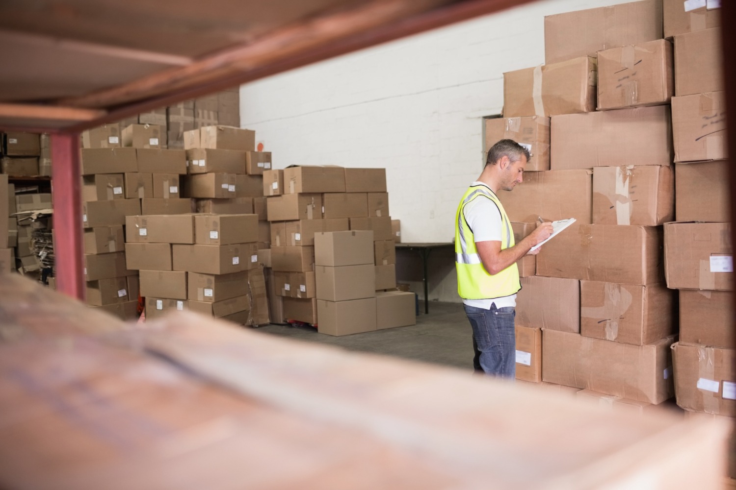 Alt text: Worker in a high-visibility vest checking inventory in a warehouse filled with stacked cardboard boxes.