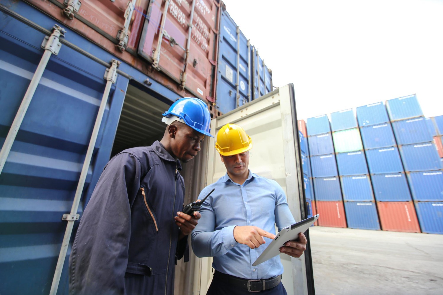 Alt text: Two logistics workers inspect a shipping container using a tablet and radio at a container yard with stacked cargo containers in the background.