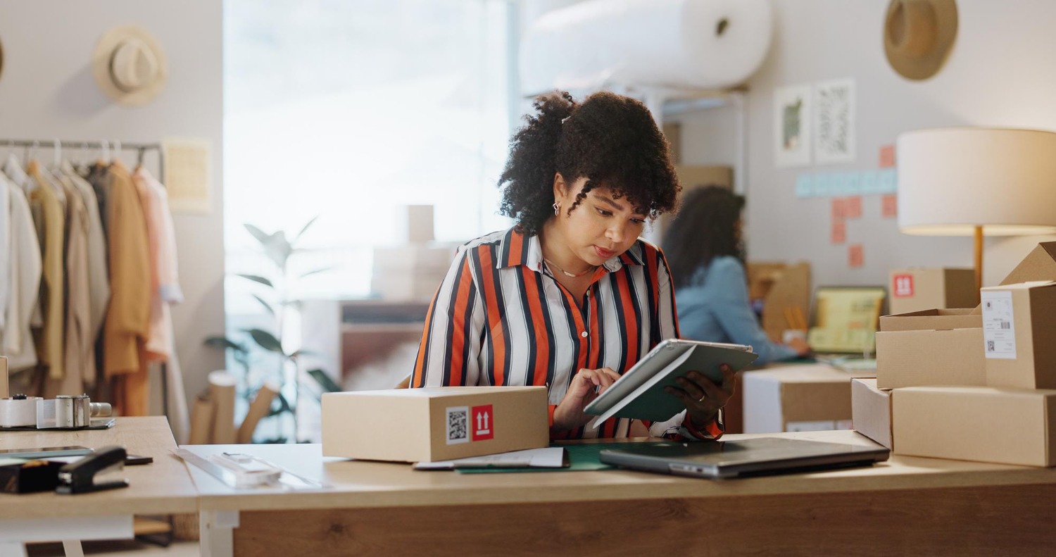 Alt text: Small business owner using a tablet to review shipping or inventory information at a packing station surrounded by boxed orders in a home or small warehouse workspace.