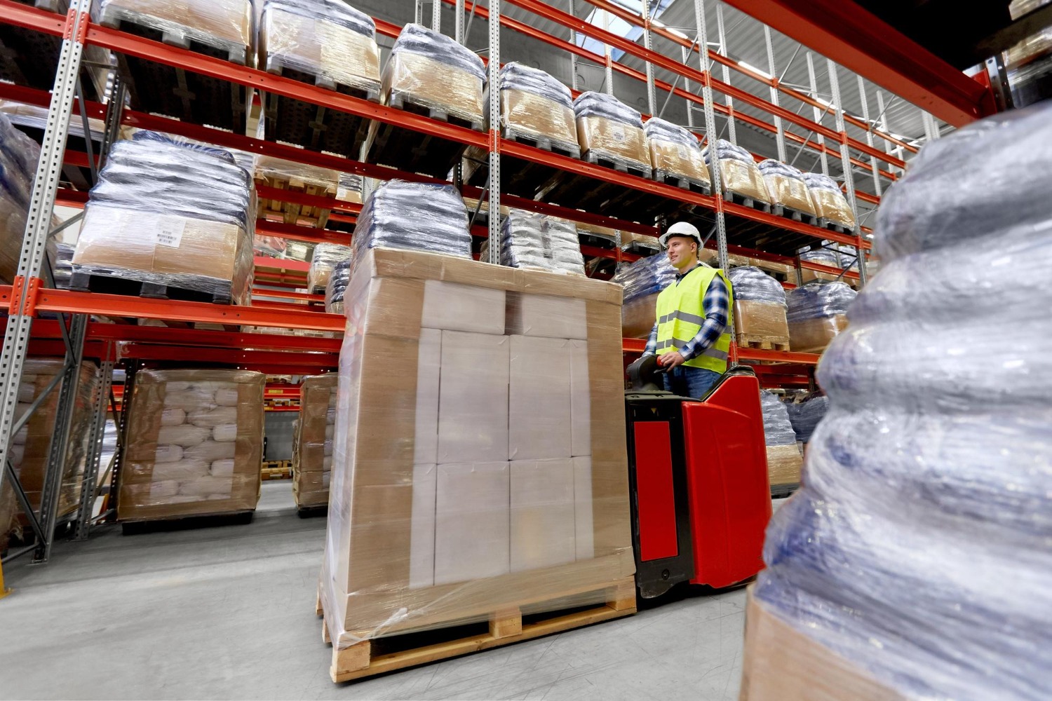 Alt text: Warehouse worker operating an electric pallet jack to transport palletised cartons through a large distribution warehouse with high-bay racking and bulk inventory storage.