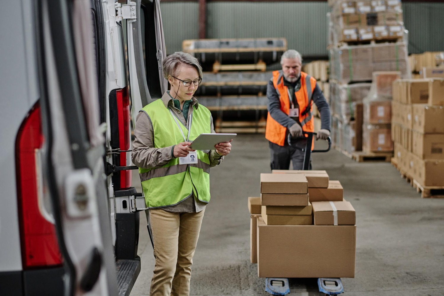Alt text: Warehouse workers preparing freight for delivery, with a logistics coordinator checking shipment details on a tablet next to a delivery van and boxed goods on a pallet jack.