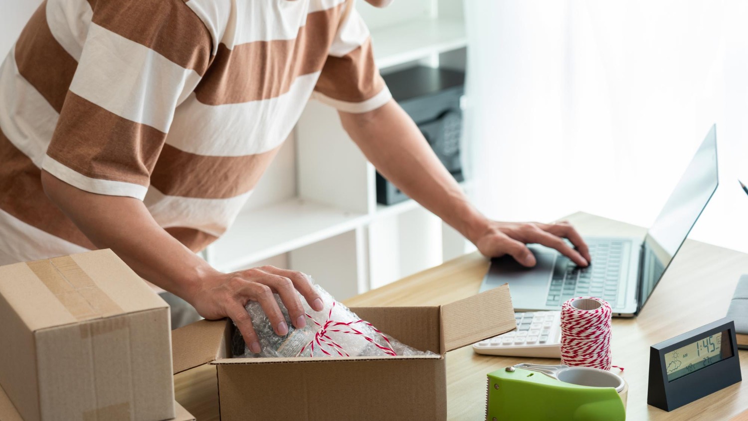 Alt text: Small online seller packing a parcel while checking orders on a laptop at a home workspace, with shipping supplies, boxes, and a calculator on the desk.