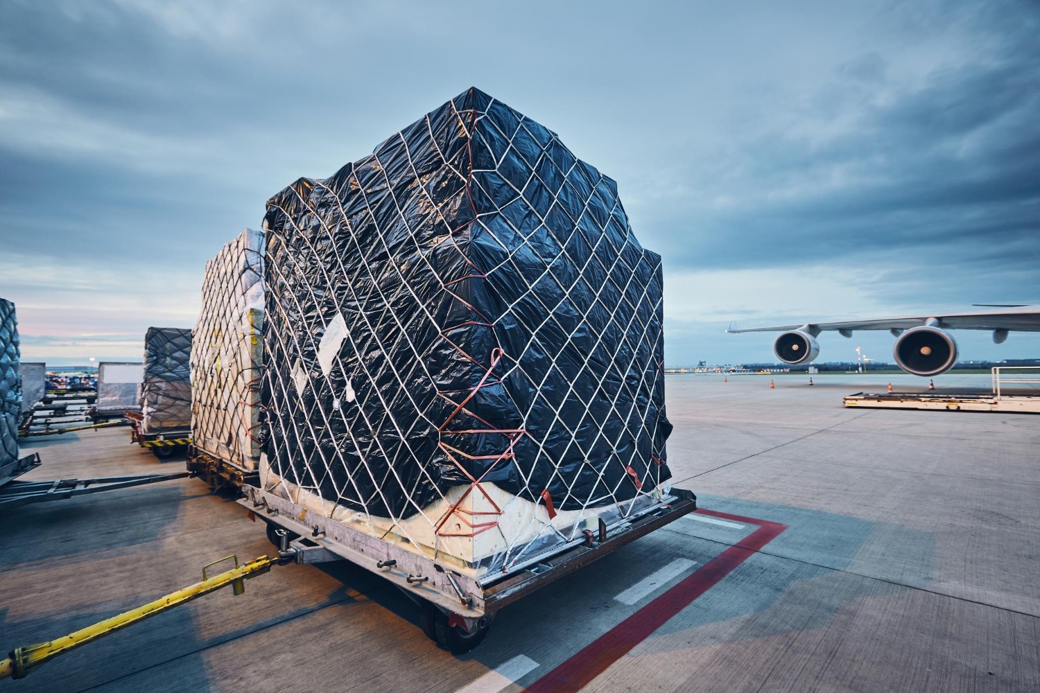 Alt text: Wrapped freight pallets on loading dollies next to a cargo plane at an international airport.