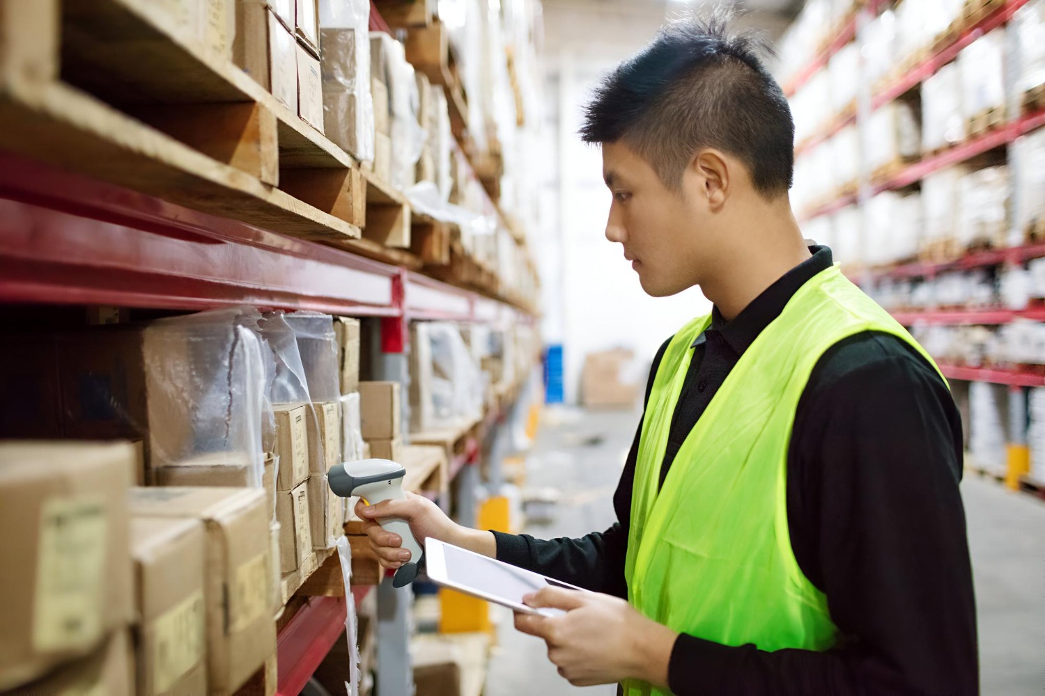 Alt text: Warehouse worker scanning inventory on warehouse shelving with a handheld barcode scanner and tablet during stock management and order fulfilment operations.