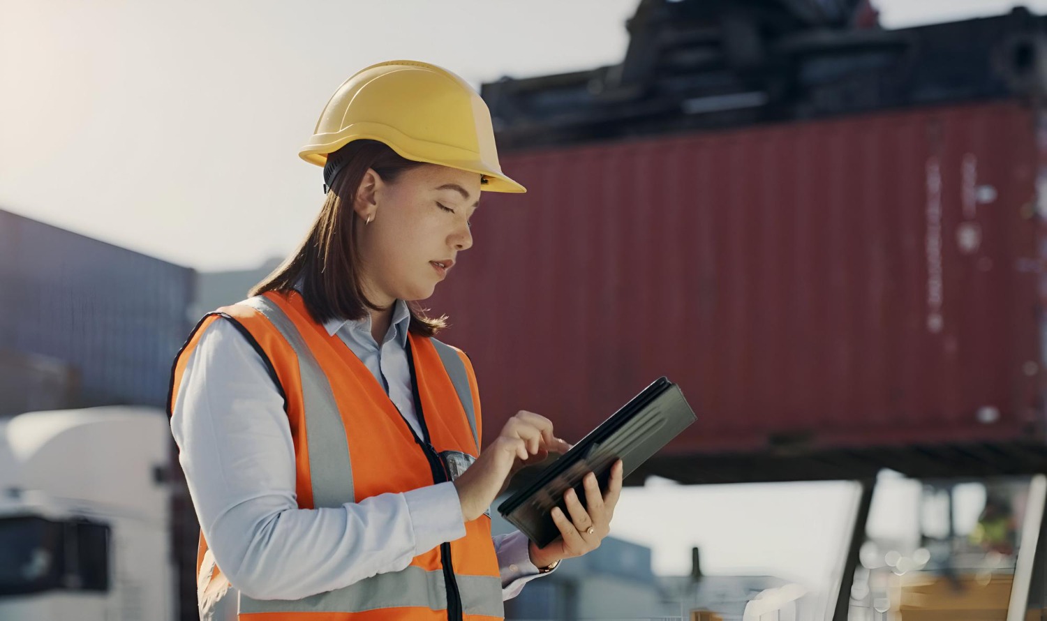 Alt text: Logistics worker in a hard hat and high-vis vest using a tablet at a container yard, with a shipping container in the background.