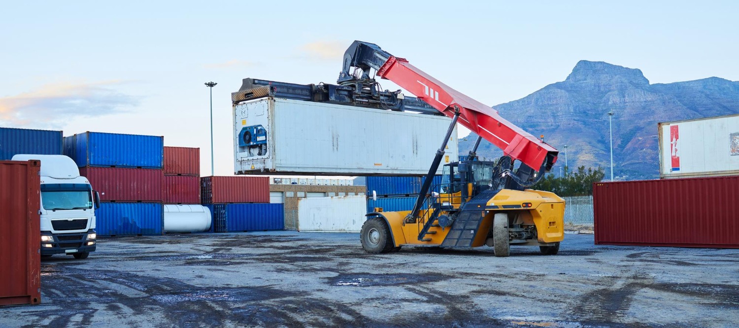 Alt text: Reach stacker lifting a shipping container in a freight yard with stacked cargo containers and a truck preparing for international freight transport.