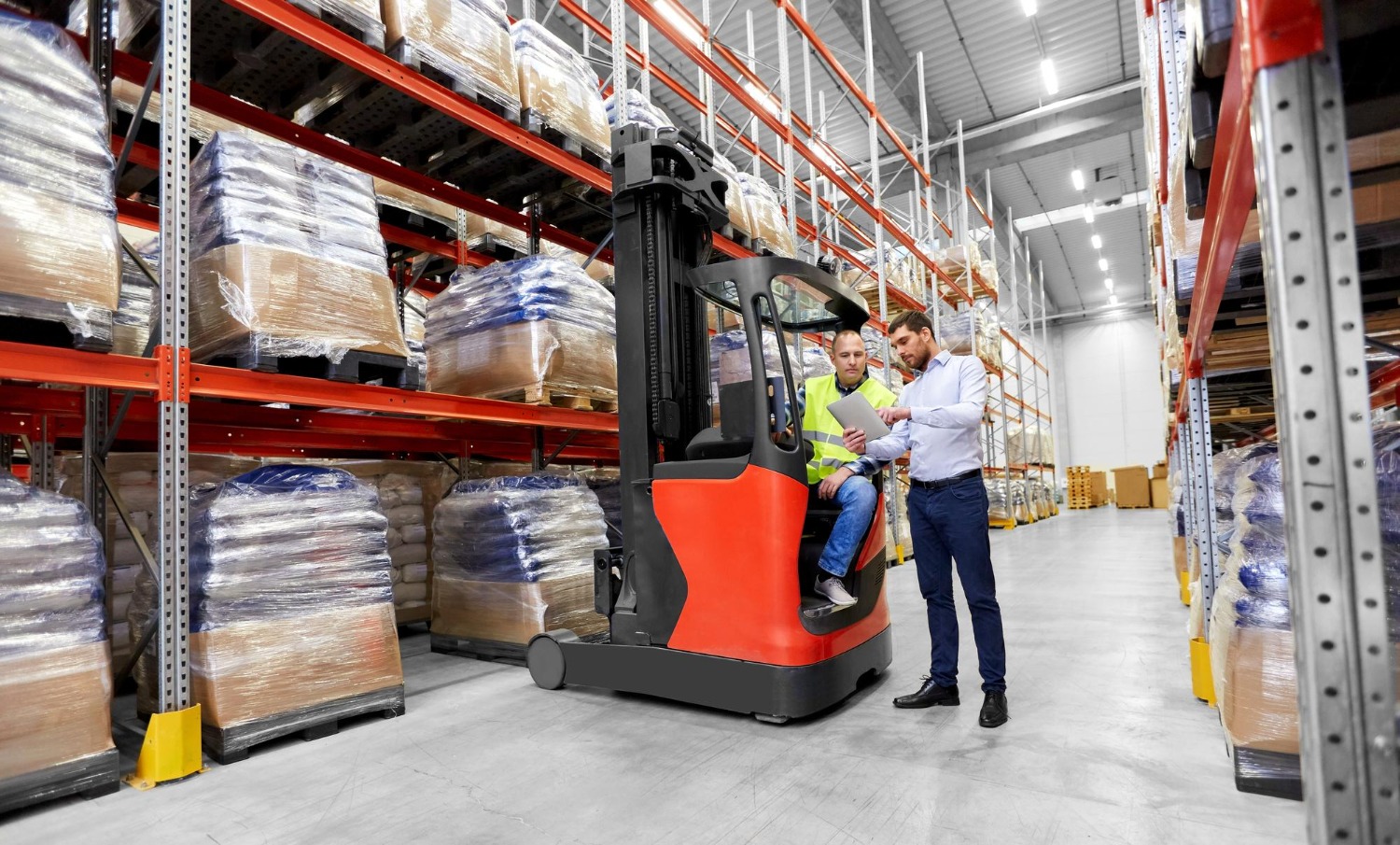 Alt text: Warehouse staff reviewing inventory beside a forklift in a pallet storage warehouse with high racking.