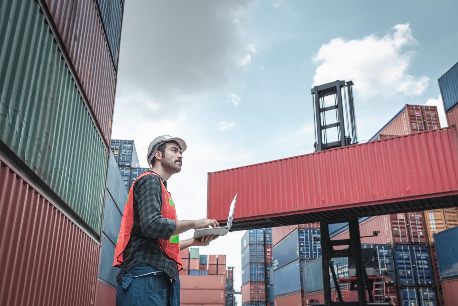 Alt text: Logistics worker using a laptop in a shipping container yard, reviewing cargo information beside stacked freight containers and a container handler.