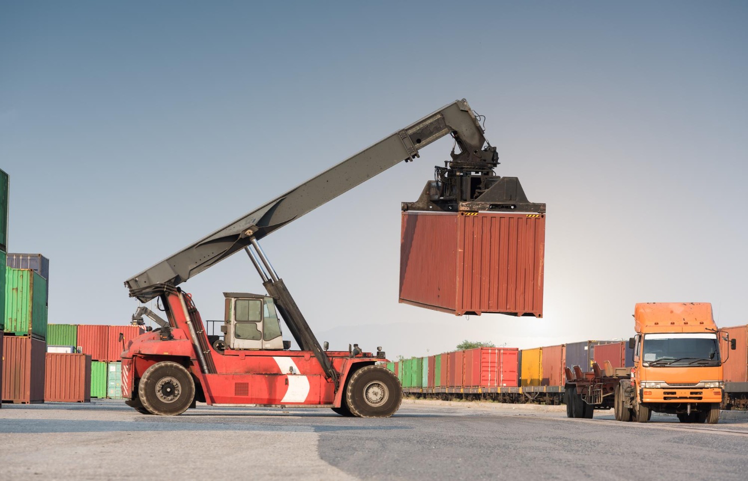 Alt text: A heavy-duty reach stacker crane lifting a red shipping container onto a freight truck at a container port terminal.