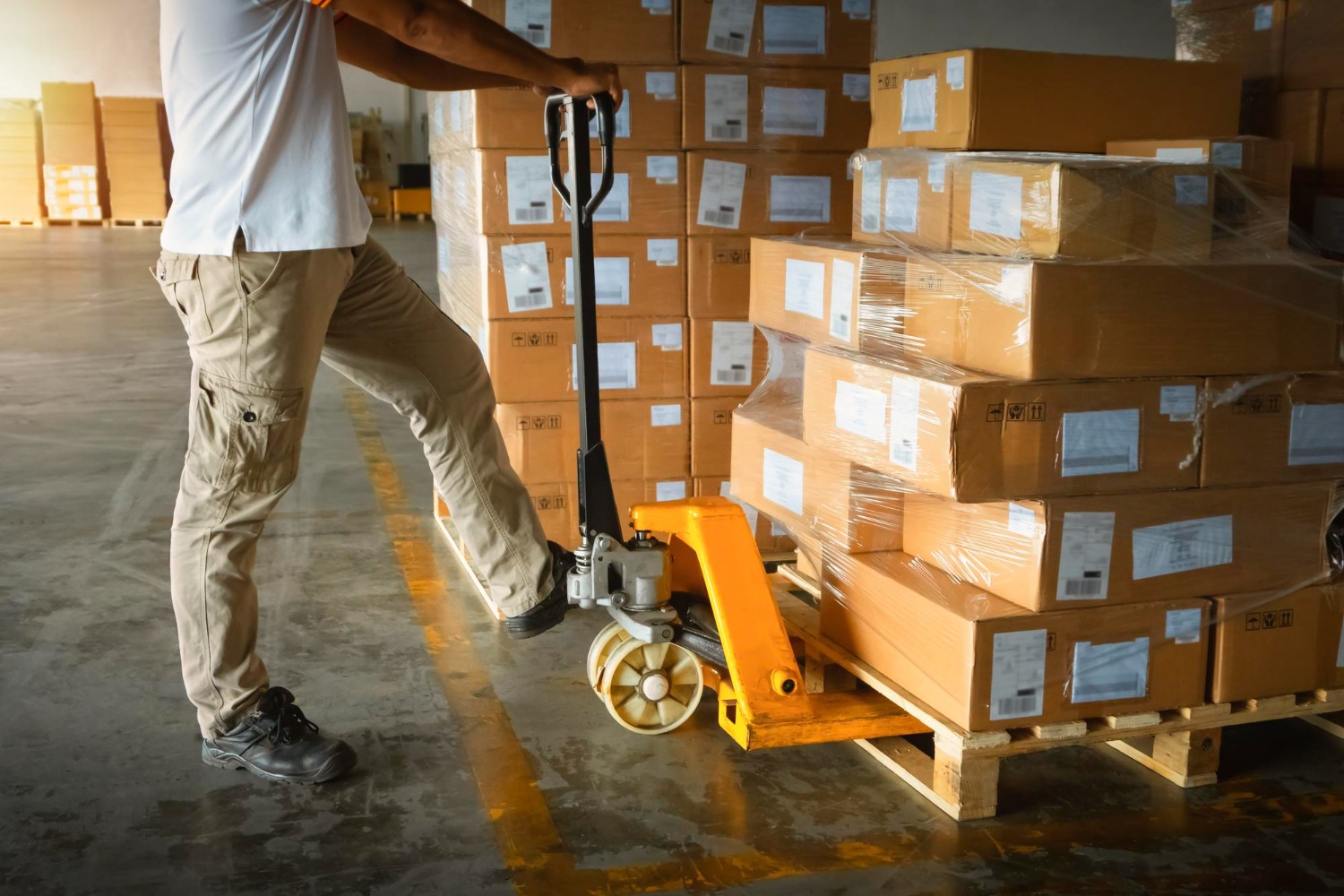 Alt text: Worker moving a pallet of boxed goods with a manual pallet jack inside a warehouse.