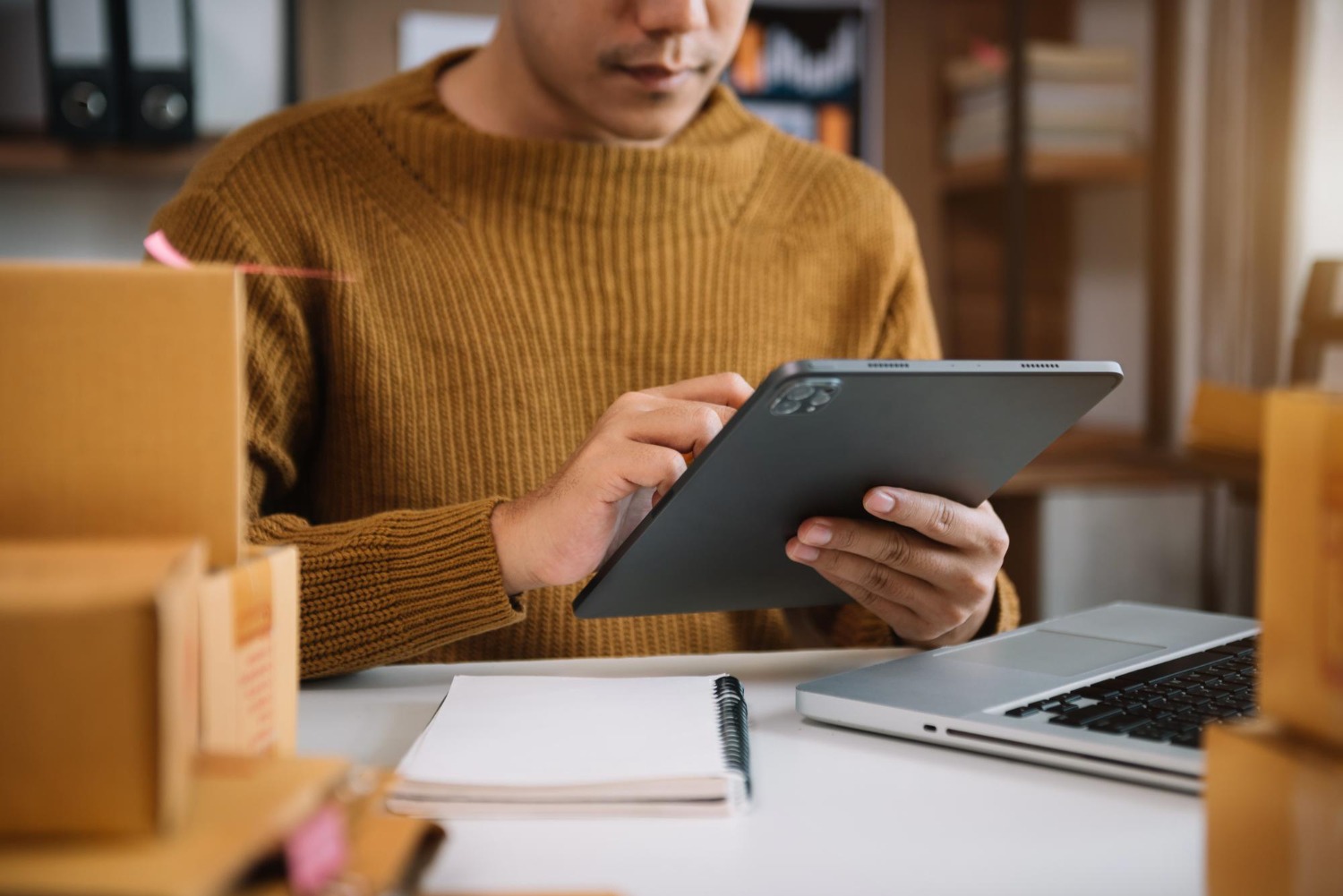 Alt text: Online seller checking inventory on a tablet beside shipping boxes and laptop.