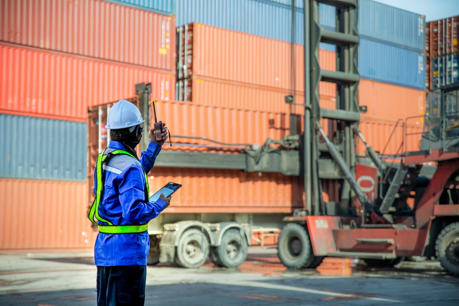Alt text: A port worker operating a container-handling vehicle at a shipping terminal, with stacked freight containers in the background.