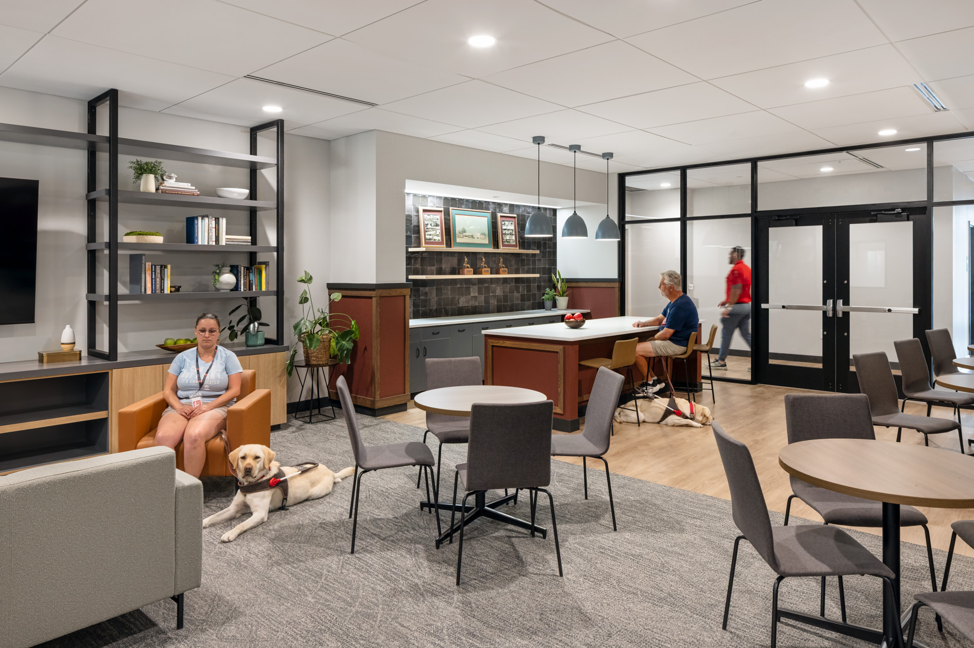 Pilot Dogs recreation room with lounge seating, café tables, a kitchenette, and students with guide dogs