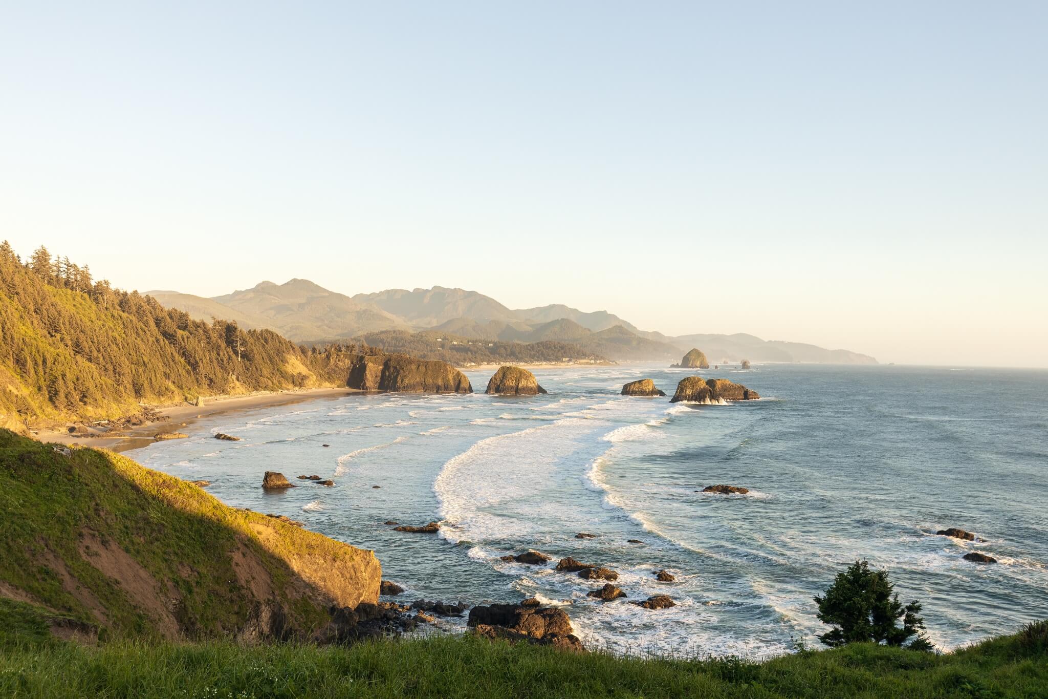 Looking up the Oregon coastline at sunset, with waves crashing into large and small rocks off the coast, and rolling hills in the distance.