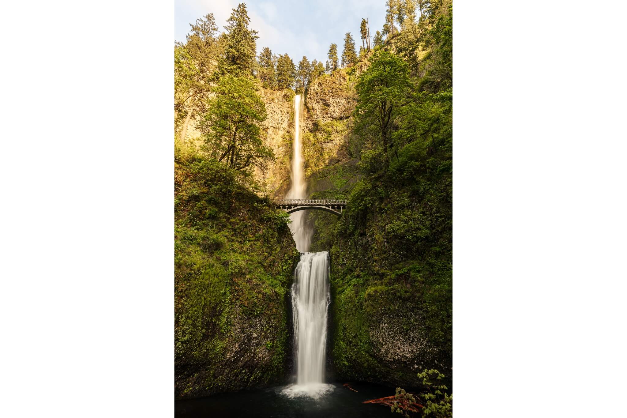 A towering set of double falls, with a historic arched bridge at the midpoint, ends in a wide pool of water. Trees and thick vegetation cover the steep cliffs on either side of the waterfall and golden sunlight lights up the top of the scene.