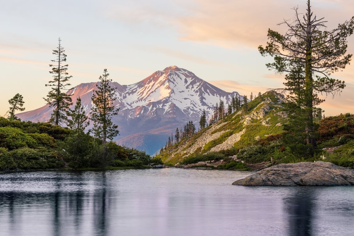 A colorful sunset with Mount Shasta lit purple and pink in the center of the image, framed on either side by small hills with evergreen trees, and a reflective lake in the foreground.