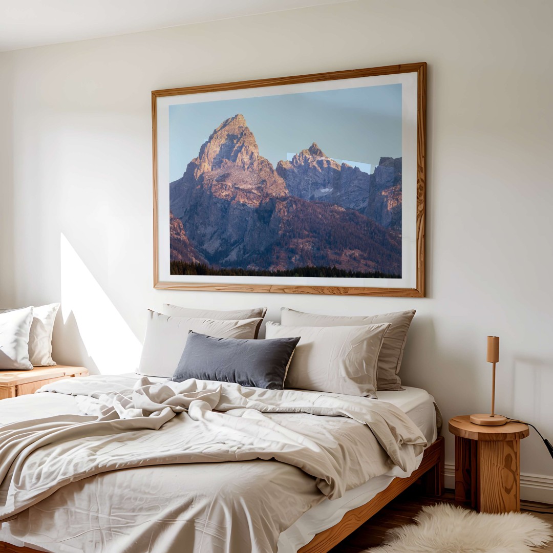 A framed photo of the Grand Teton mountain peak at sunrise hanging on a wall above a bed
