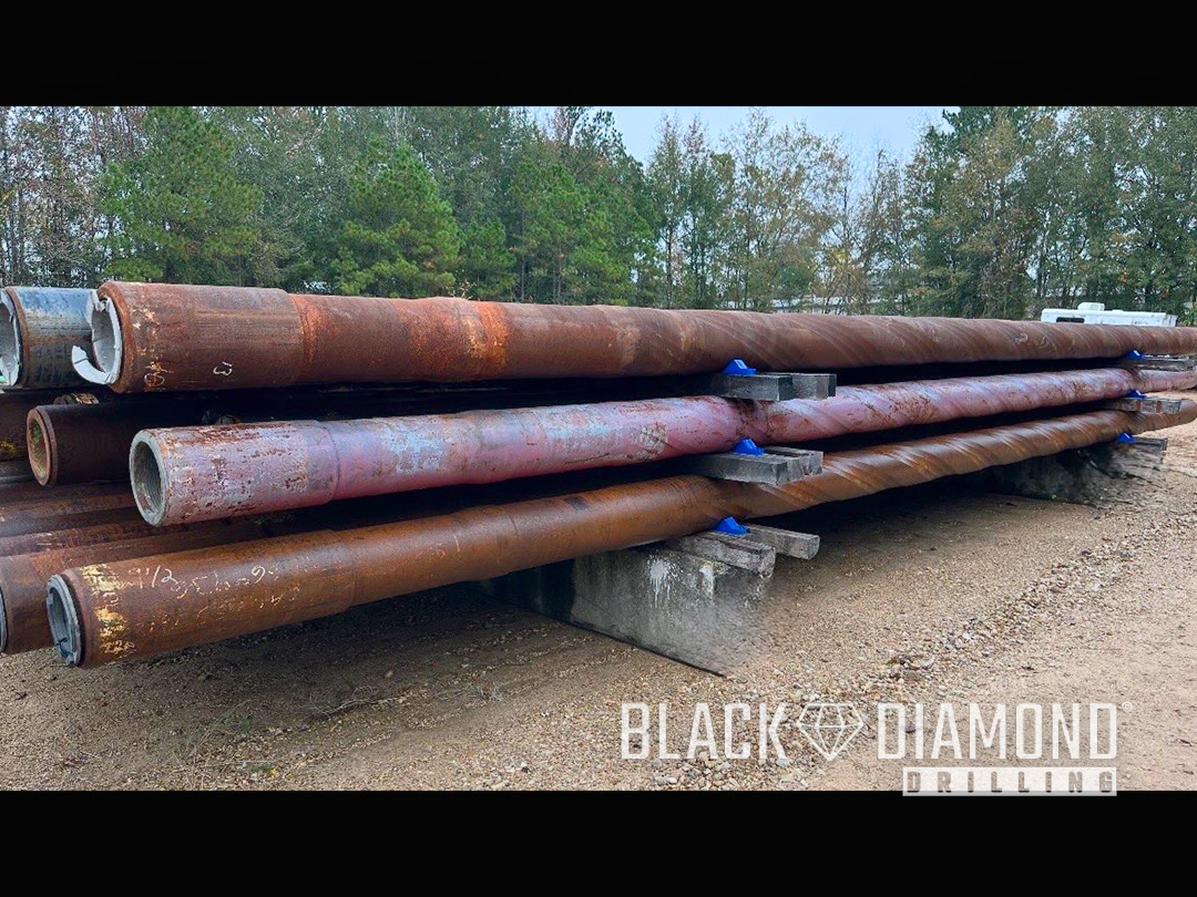 A stack of 9-1/2 inch spiral drill collars with 7-5/8 REG connections stored on a rack in an outdoor pipe yard.