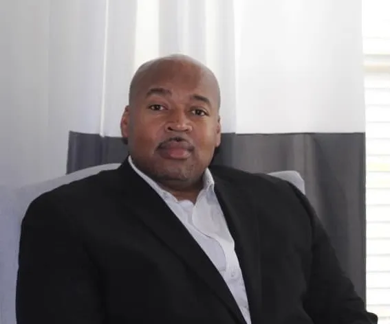 Man in a black suit jacket and white shirt seated in front of curtains.