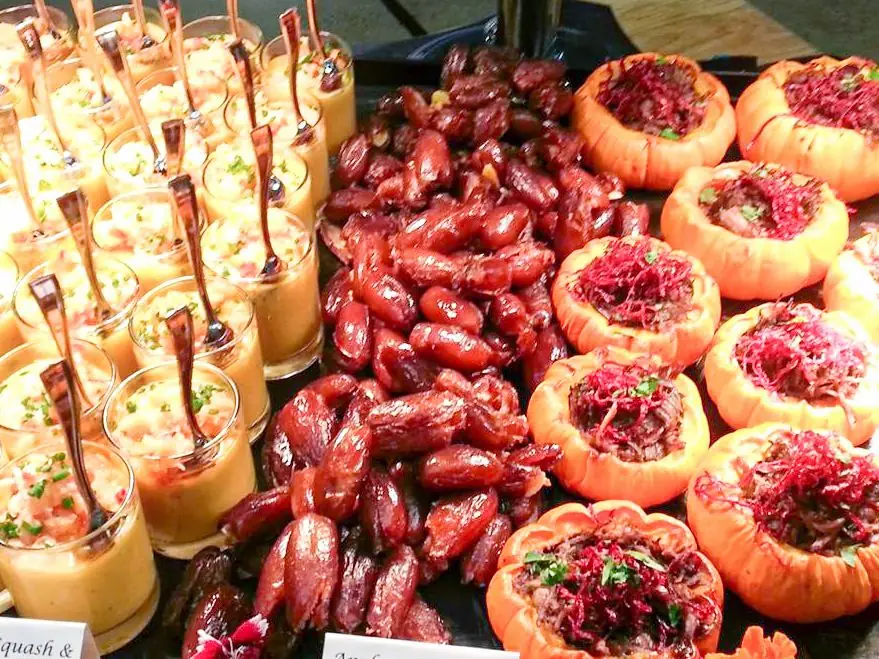 Tray of small glasses filled with creamy squash soup garnished with herbs, glazed cocktail sausages, and hollowed mini pumpkins stuffed with savory mixture and topped with shredded red garnish.