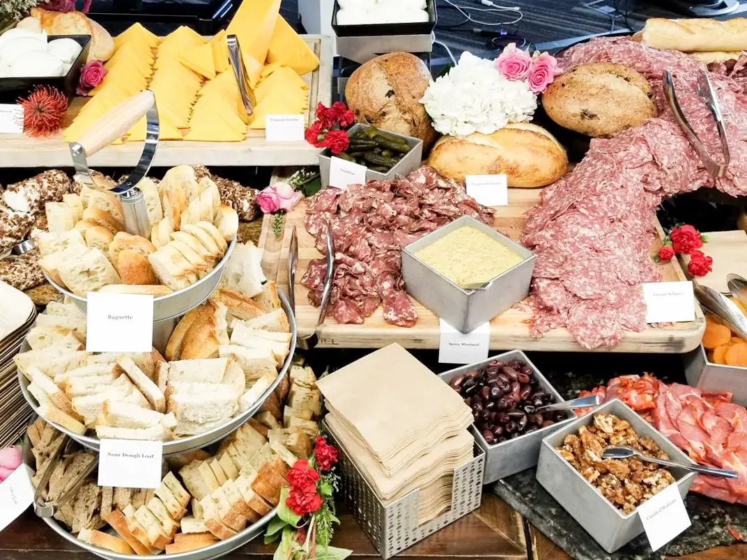 Buffet with assorted sliced breads, cheddar cheese, cured meats, pickles, olives, nuts, and dipping sauces presented on wooden boards and metal trays with floral decorations.