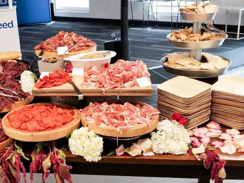 Charcuterie spread with various sliced cured meats, bowls of dip, stacked plates, crackers, and decorative white and red flowers.