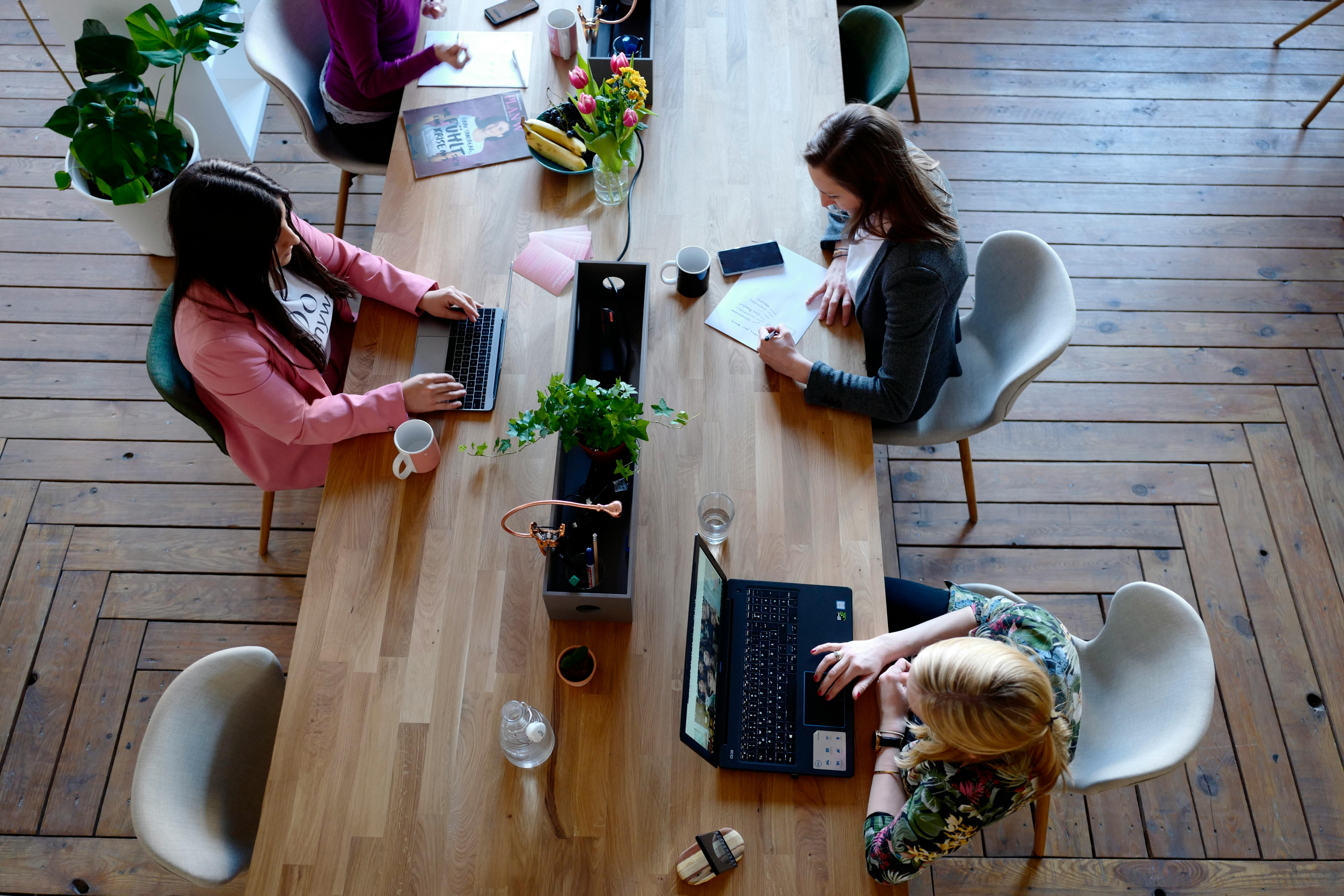 Overhead view of people working at a shared wooden table with laptops and notebooks