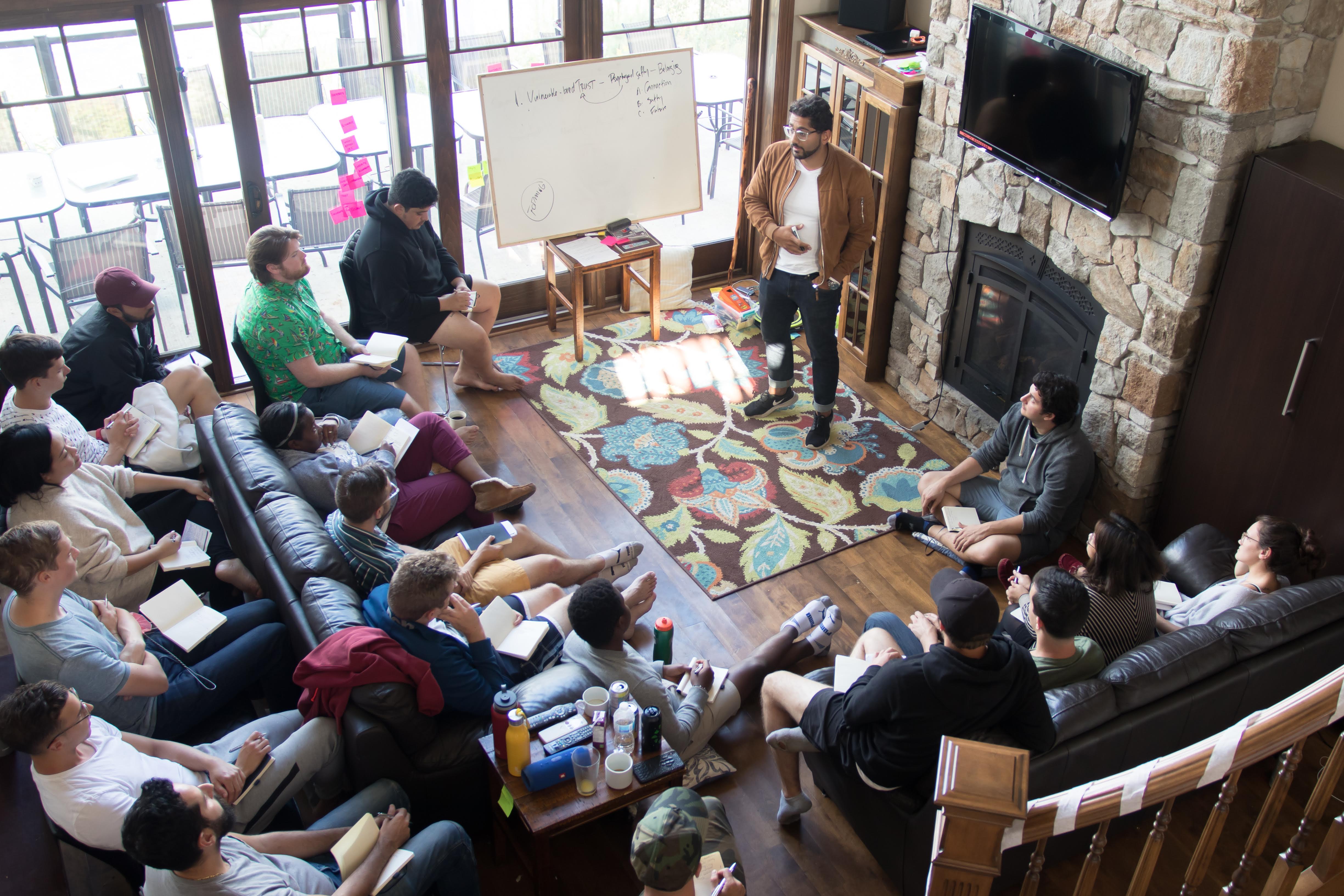 A facilitator stands in a cozy living room leading a small group workshop. Participants sit on couches and the floor in a circle, taking notes while listening. A whiteboard with handwritten notes stands near large windows, and a stone fireplace and mounted TV frame the space, creating an intimate team retreat setting.