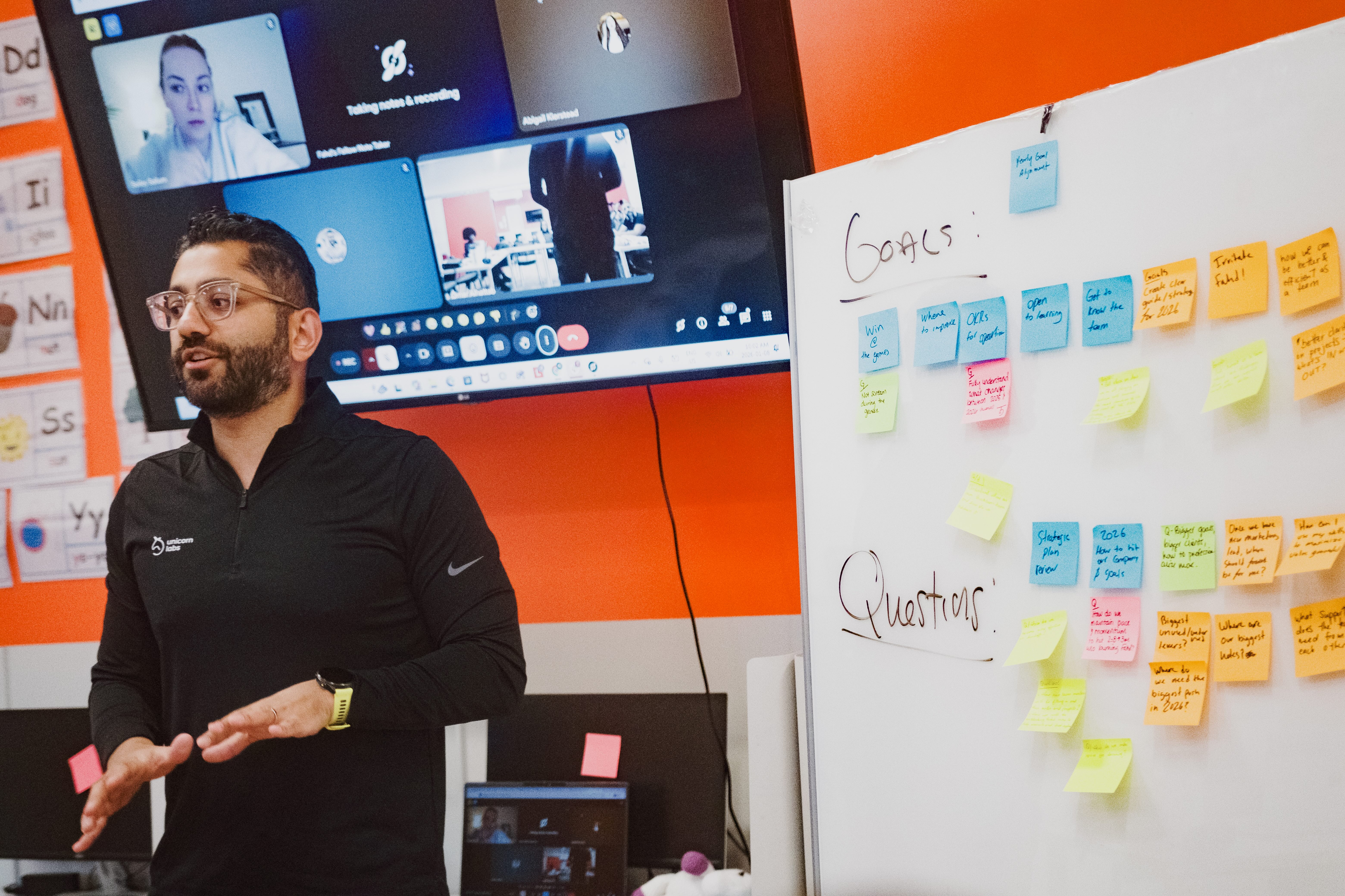 Fahd in a black quarter-zip presenting beside a whiteboard covered in colourful sticky notes labeled “Goals” and “Questions,” with a virtual meeting displayed on a screen behind him in an orange-walled room.