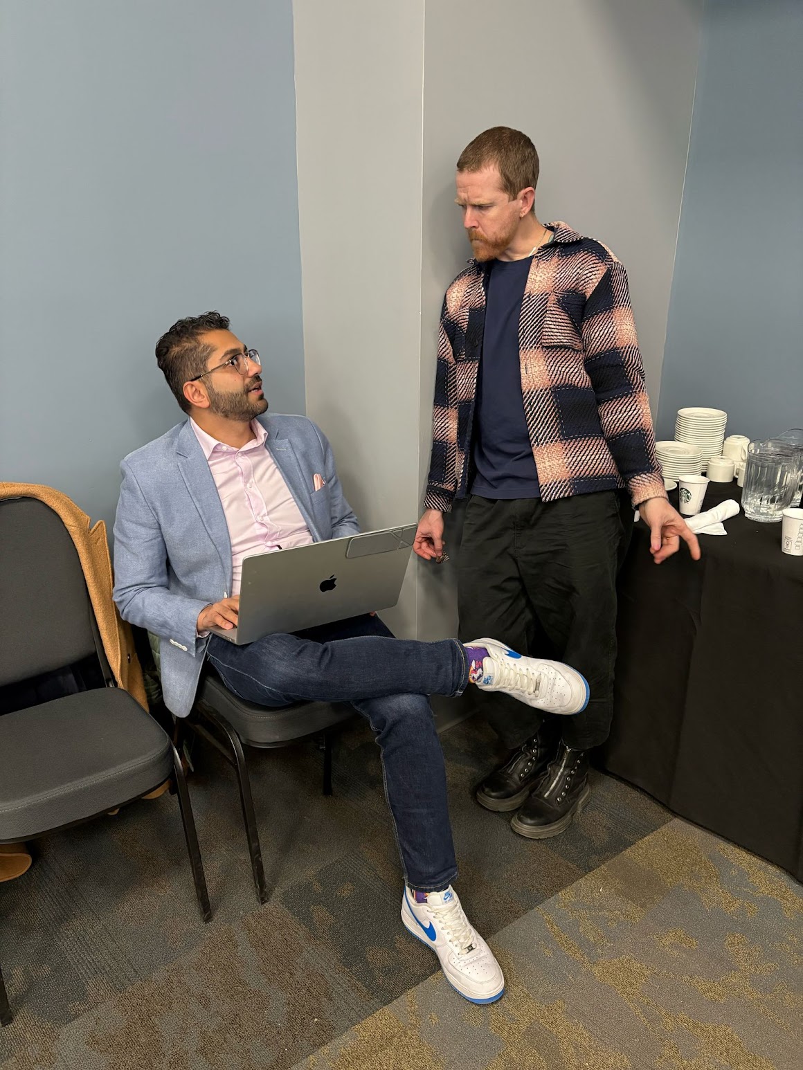 Two men having a conversation backstage at an event, one seated with a laptop.