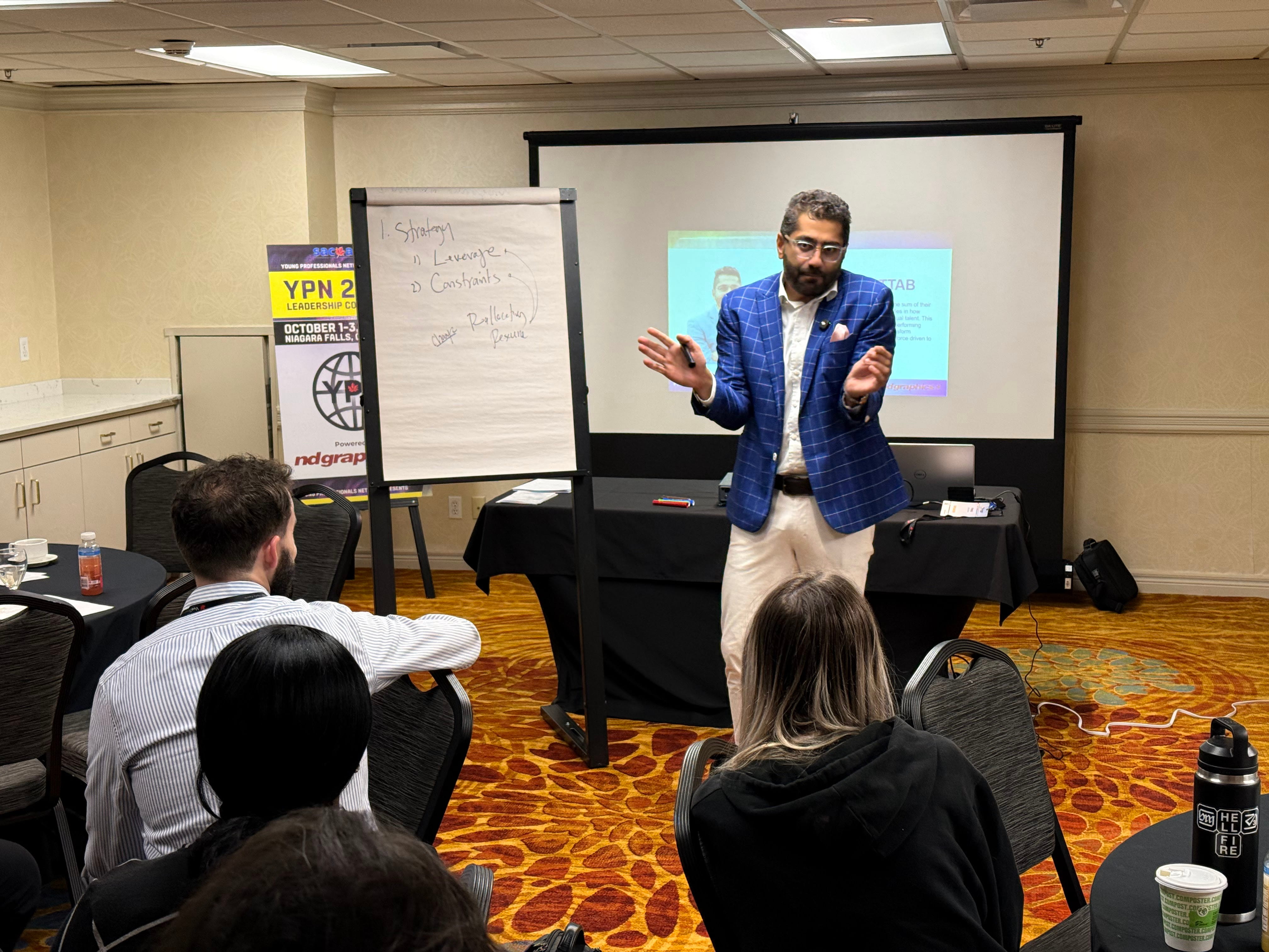 Fahd Alhattab facilitating a workshop at a YPN Leadership Conference in Niagara Falls, wearing a blue plaid blazer, gesturing with both hands in front of a flipchart with strategy notes.