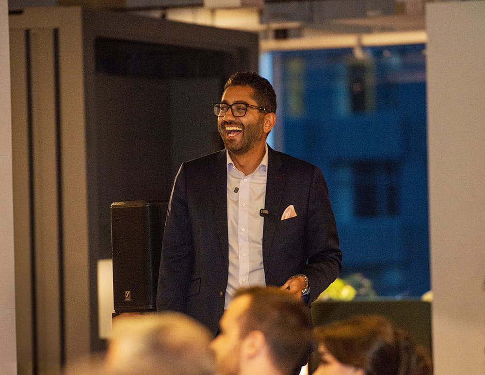  Fahd Alhattab smiling broadly while presenting in a dimly lit venue, wearing a navy suit and pink pocket square, with an audience visible in the foreground.