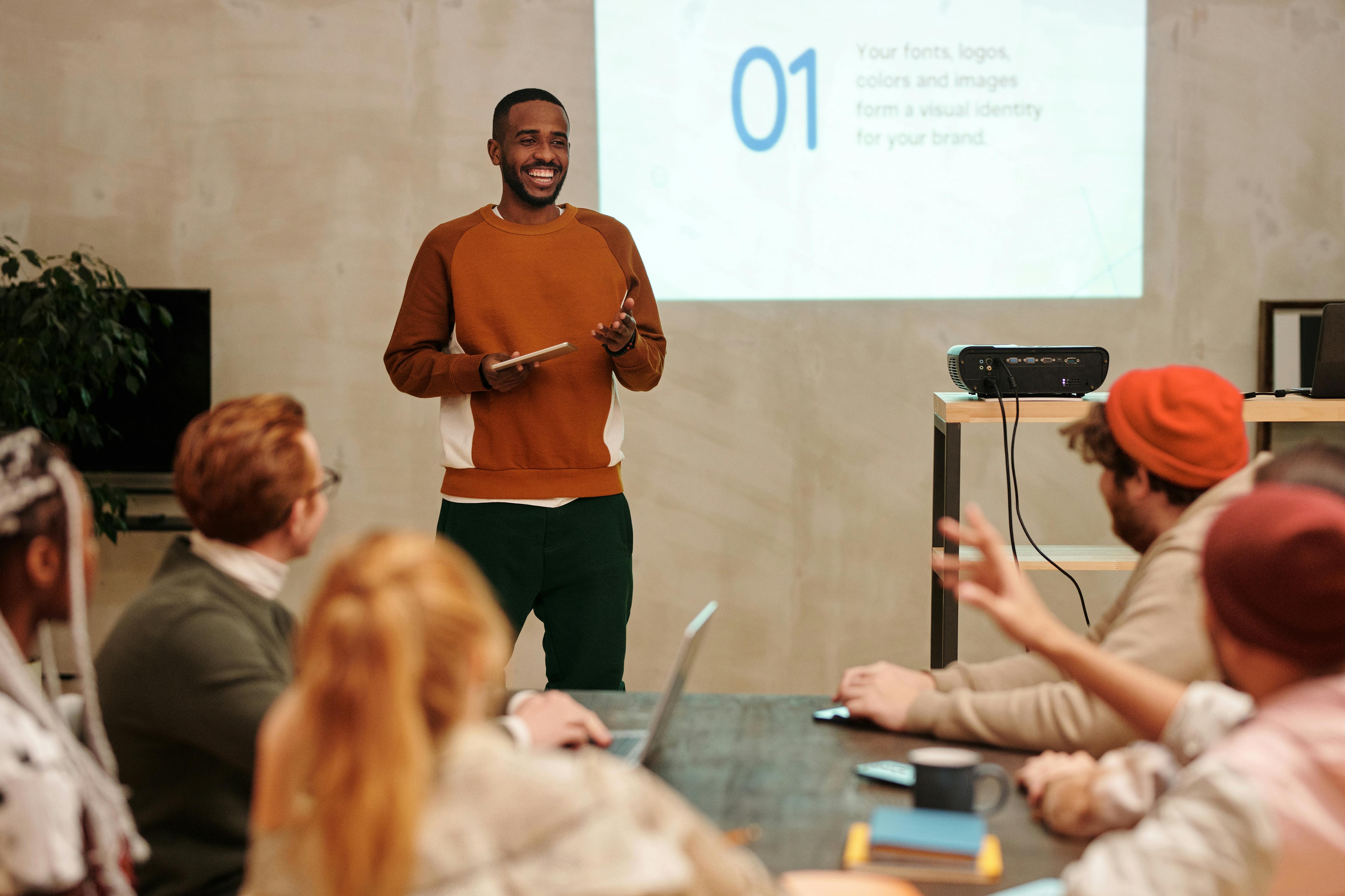 A smiling man in an orange sweater presenting to an engaged, diverse seated audience, holding a tablet, with a projected slide showing brand identity content behind him.