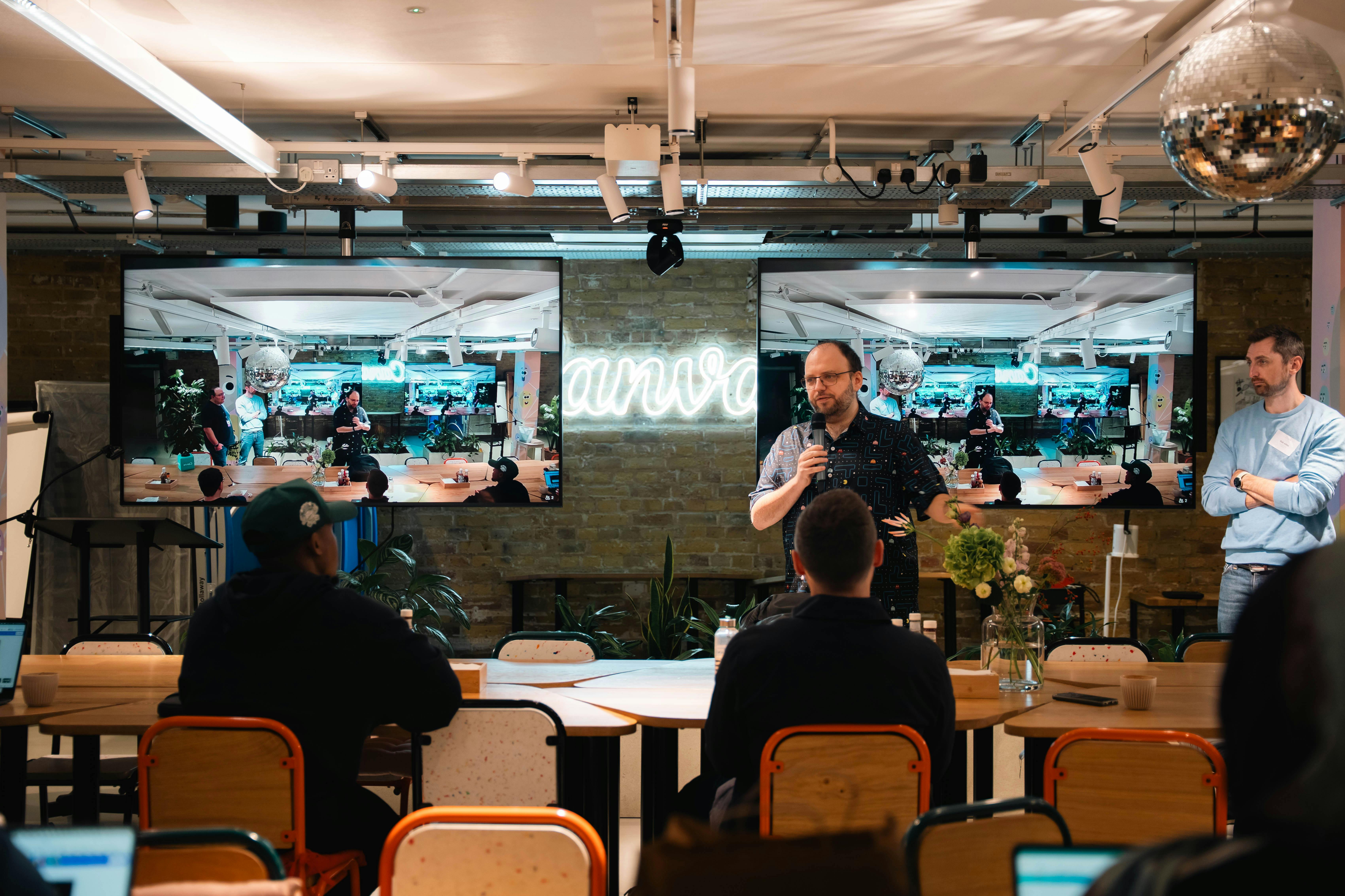 A man in a patterned shirt speaking at a Canva-branded event space with a disco ball overhead, two large screens mirroring the room behind him, and a second presenter standing to the side.