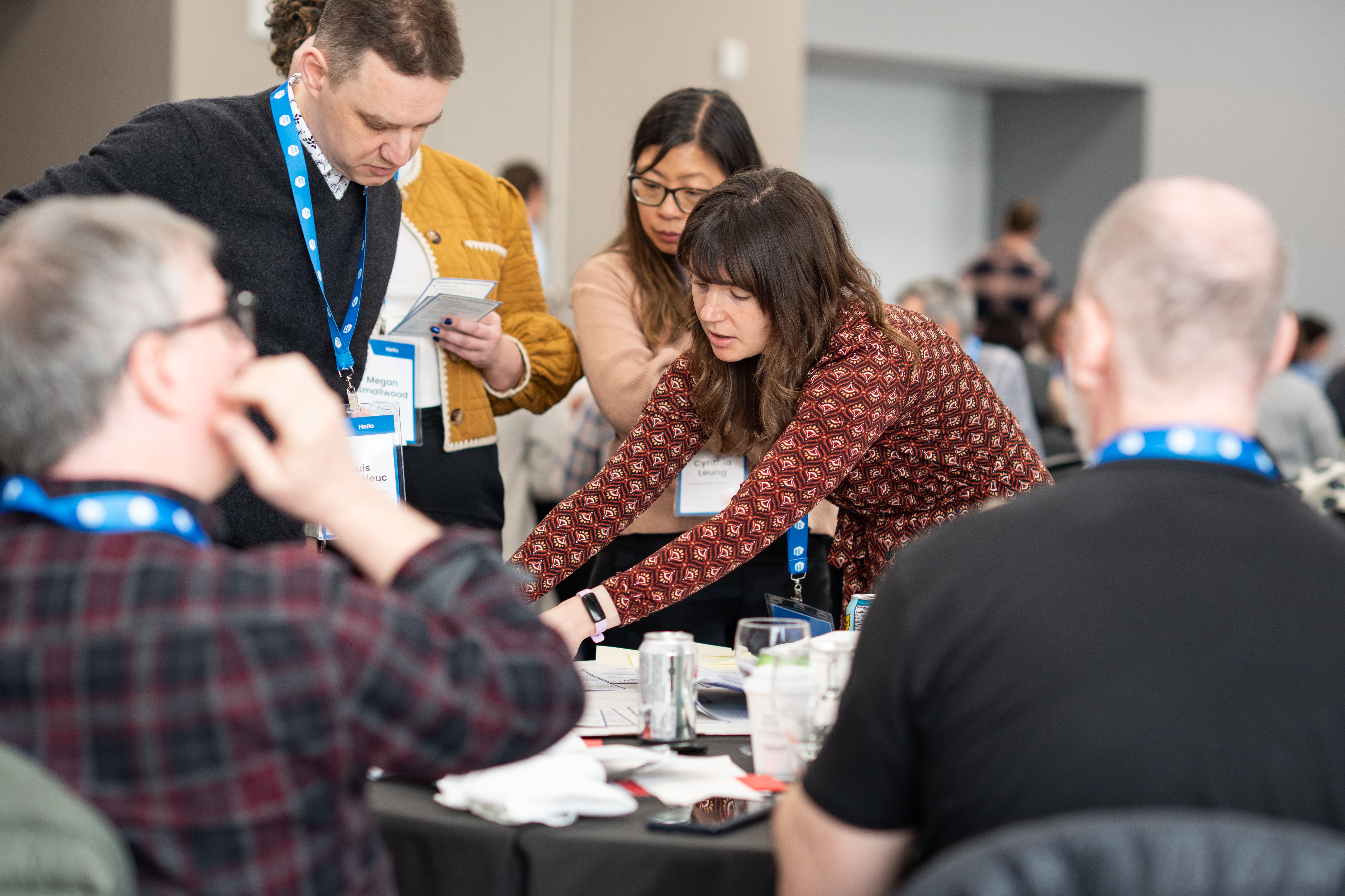 Participants at a leadership workshop collaborating around a table during a group exercise, reviewing worksheets and discussing ideas with name-badge lanyards on