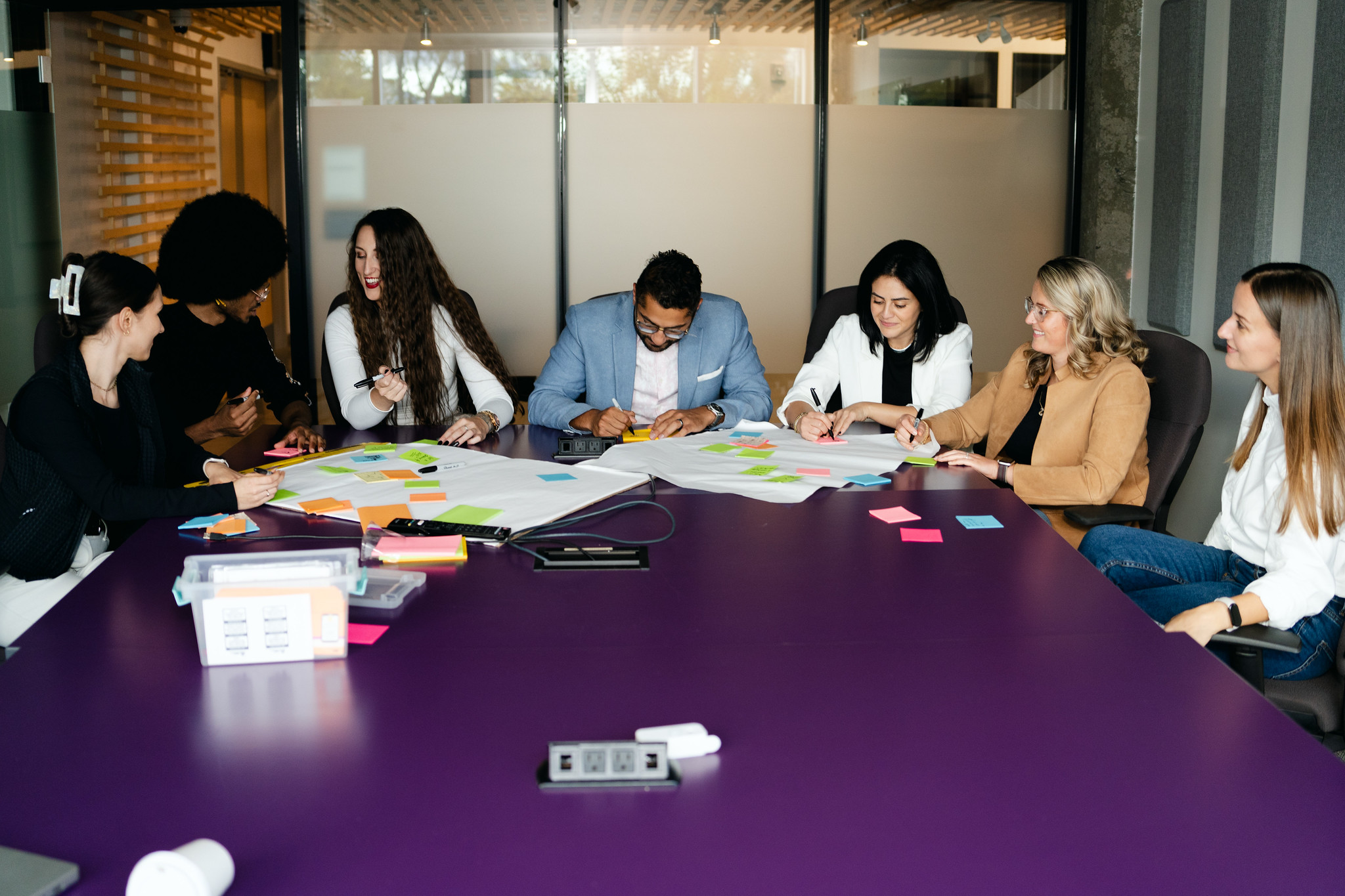 Diverse team gathered around a conference table during a facilitated workshop, using colourful sticky notes and markers on large paper for a group brainstorming exercise