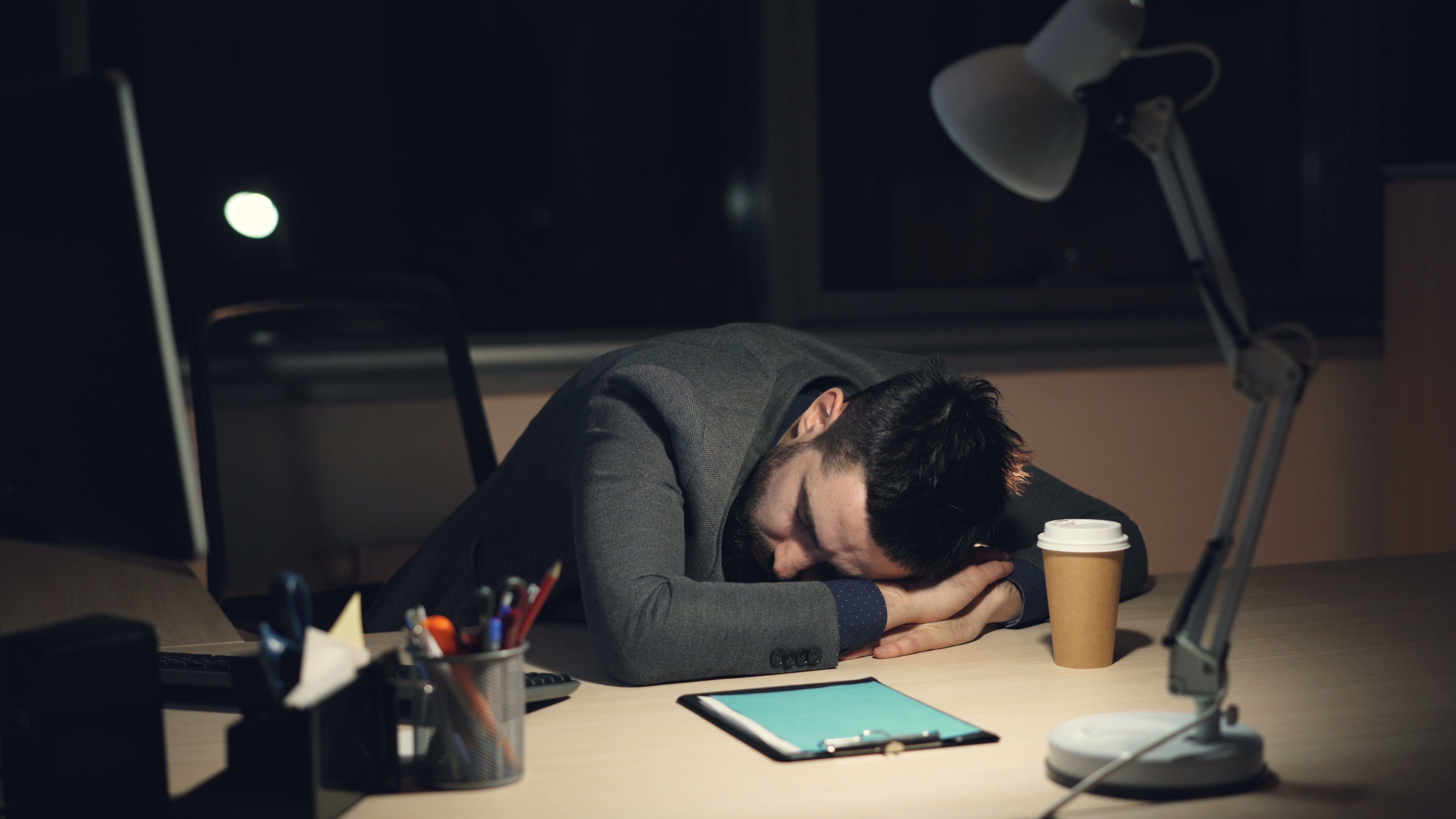Exhausted manager asleep at his desk late at night beside a laptop and coffee cup, illustrating burnout and overwork
