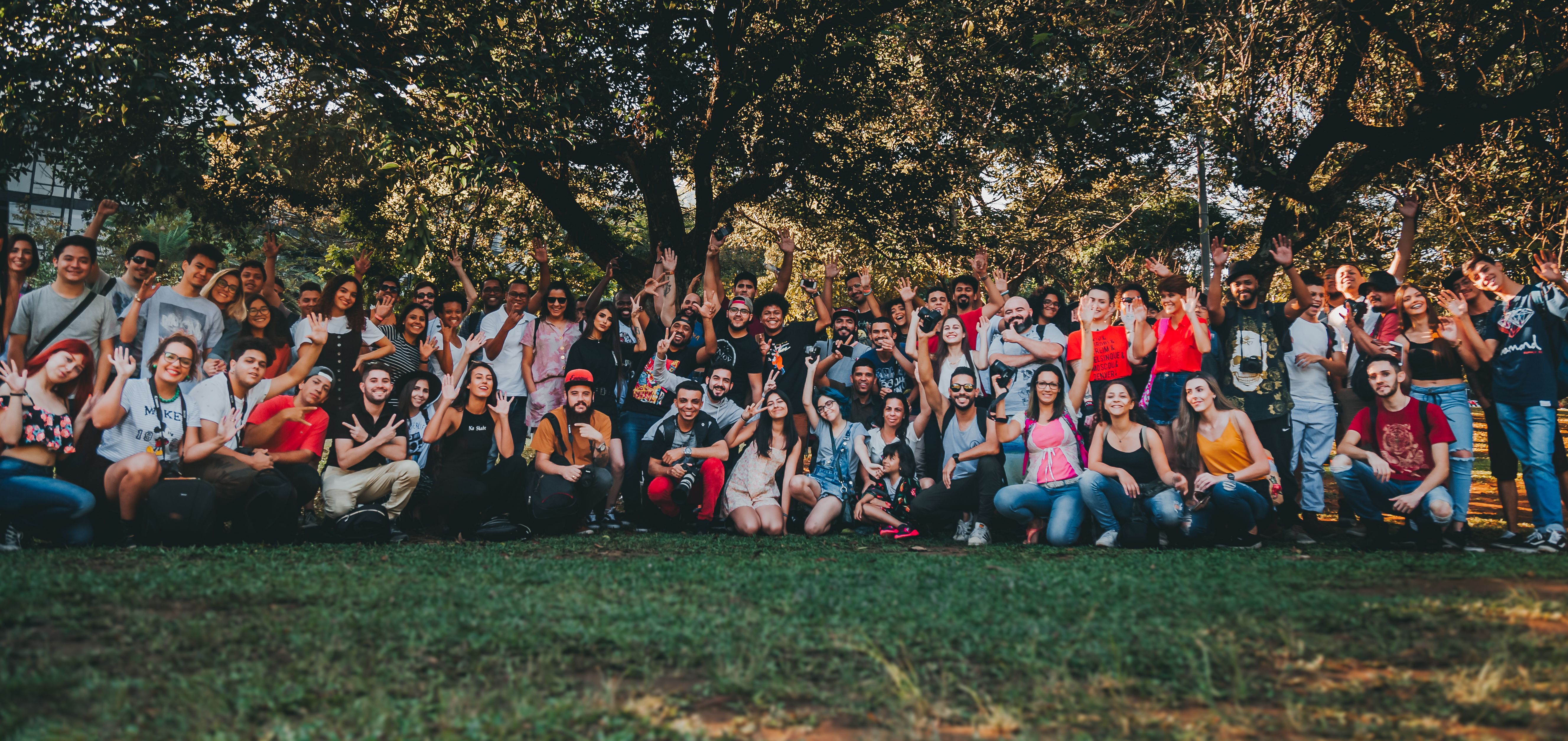 Large group of people posing together outdoors under a tree, smiling and waving in a big team or community photo