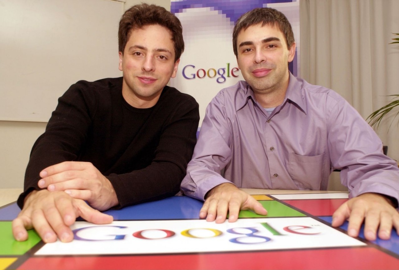Google co-founders Sergey Brin and Larry Page seated behind an early multicolour Google logo in an office