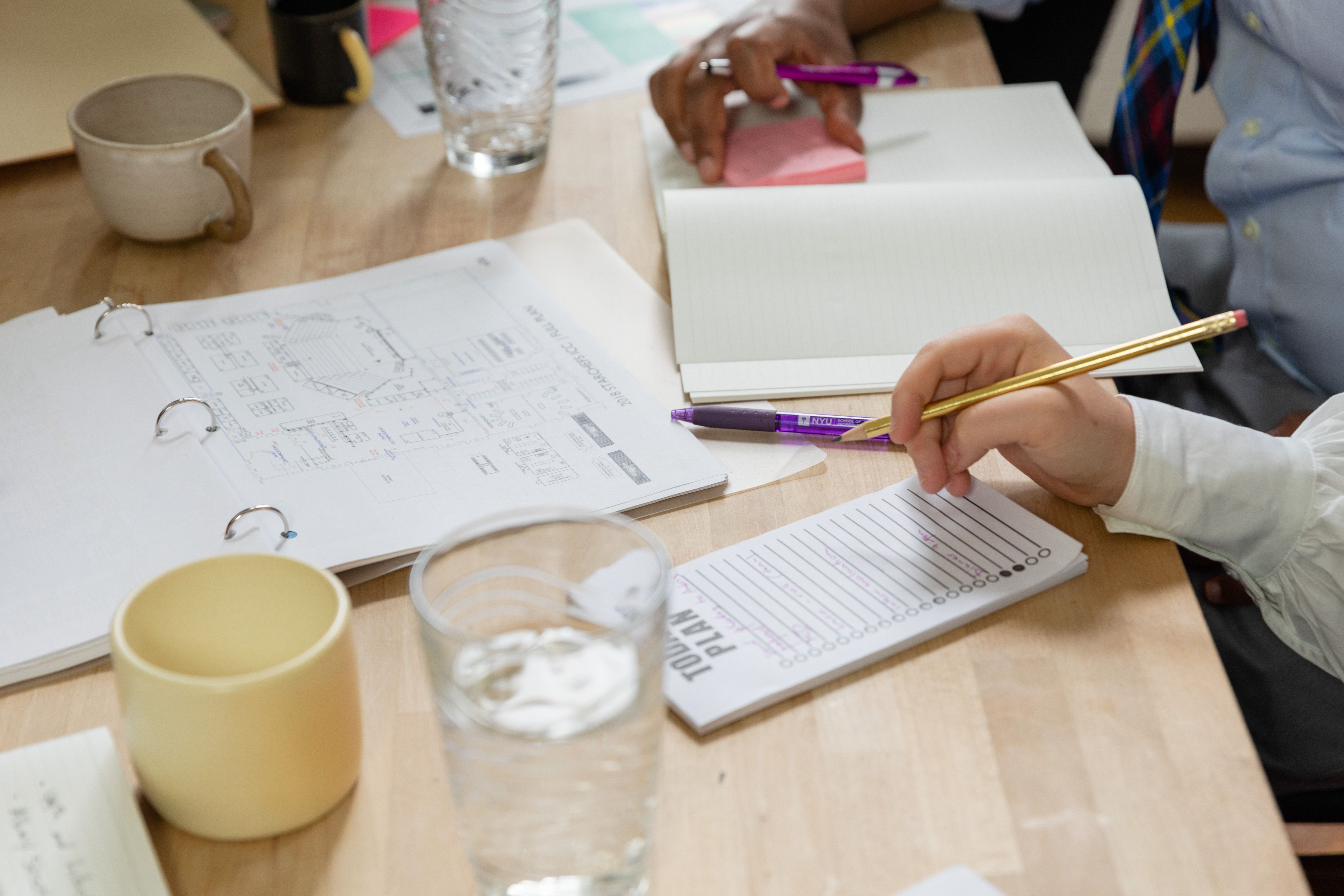Close-up of workshop participants planning at a table with an open binder, floor-plan document, notepads, and a printed action plan worksheet