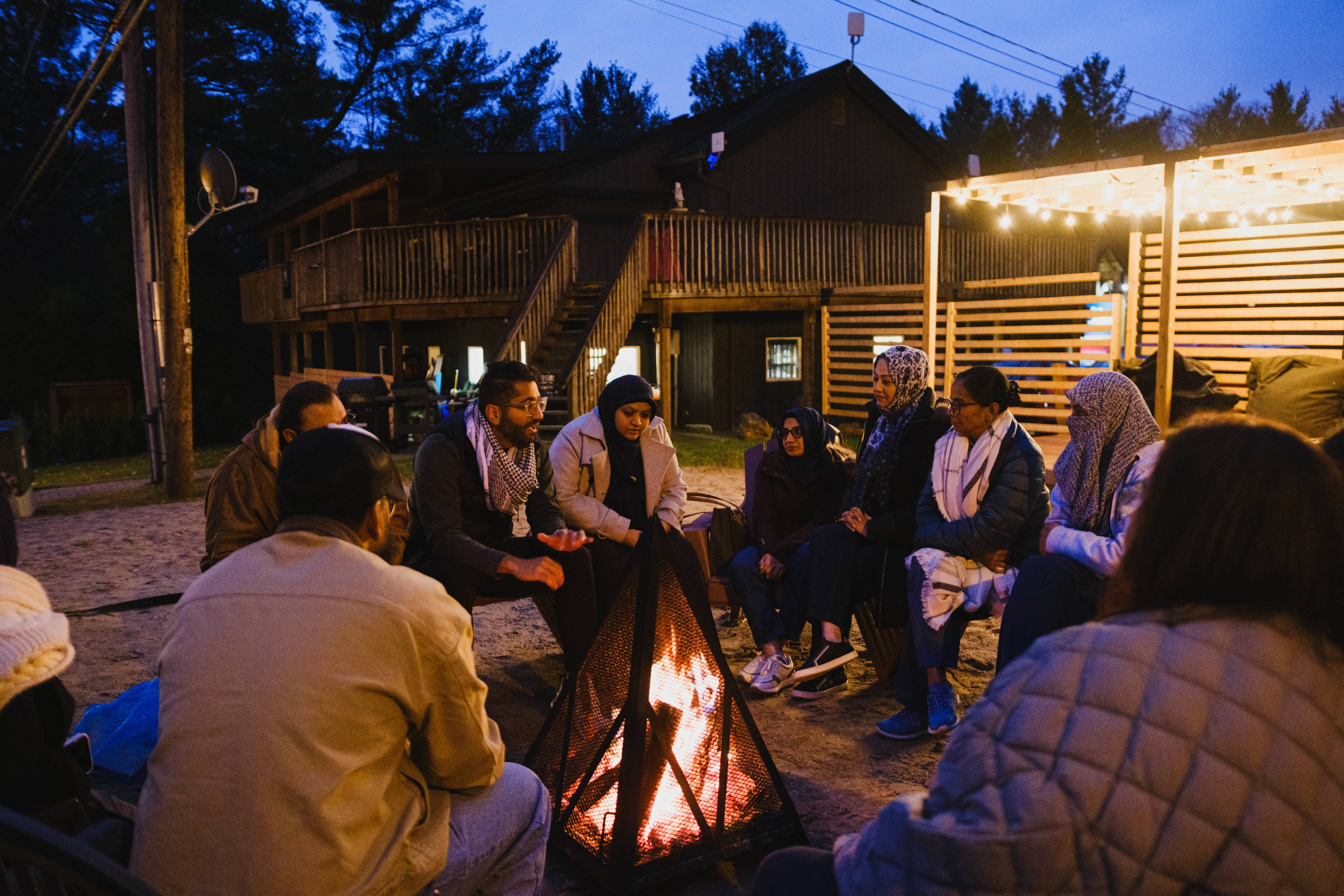 Group gathered around an outdoor fire pit at dusk in front of a wooded lodge, having a reflective conversation during a leadership retreat