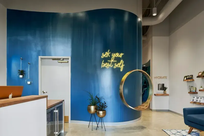 Lobby at sēk Sauna Studio with a blue wall, a gold ring swing and a neon sign that reads sēk your best self