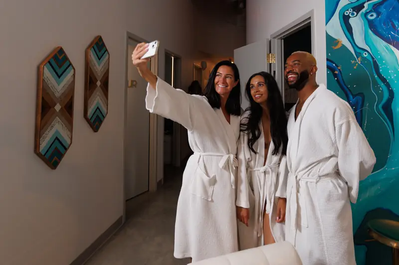 Three friends in robes taking a selfie in the hallway at sēk Sauna Studio, beside the geode mural