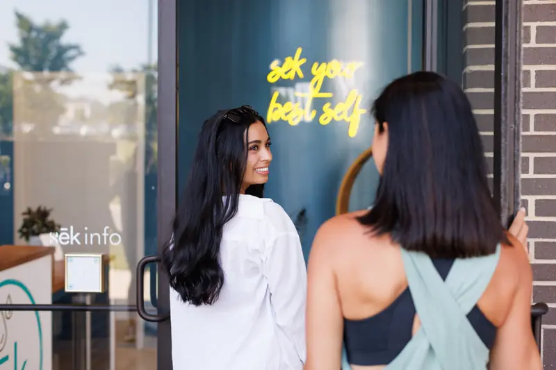 Two friends arriving at the front door of sēk Sauna Studio in Franklin, TN, under a neon sign reading sek your best self