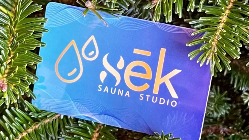 A physical sēk Sauna Studio gift card in blue and gold, resting on evergreen branches