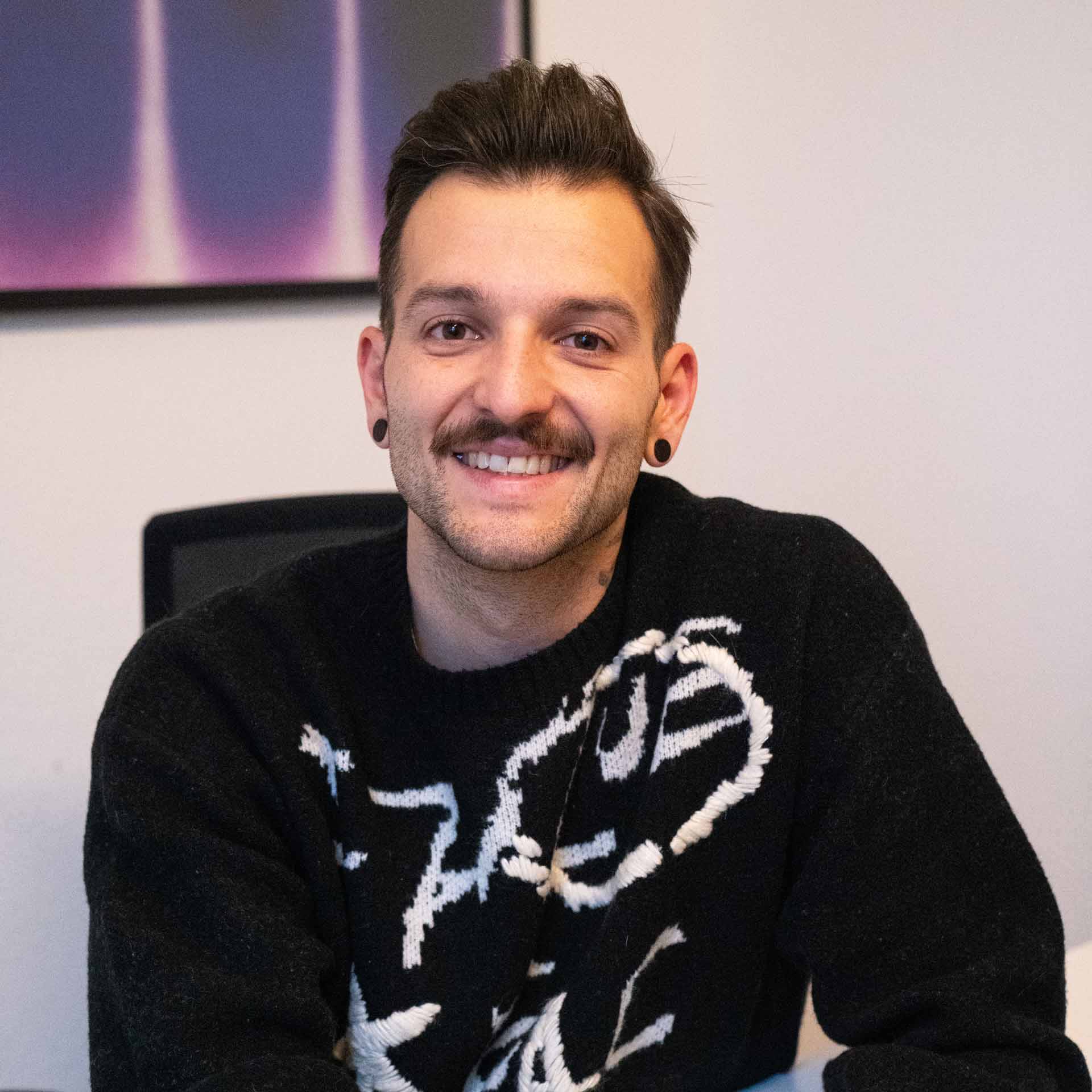 Junger Mann mit Schnurrbart, schwarzen Ohrsteckern und einem schwarzen Pullover sitzt lächelnd vor hellem Hintergrund.