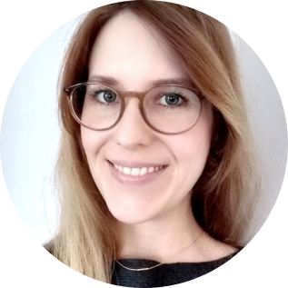 Porträt einer lächelnden Frau mit langen, hellbraunen Haaren und braunen Brillengestellen vor weißem Hintergrund.