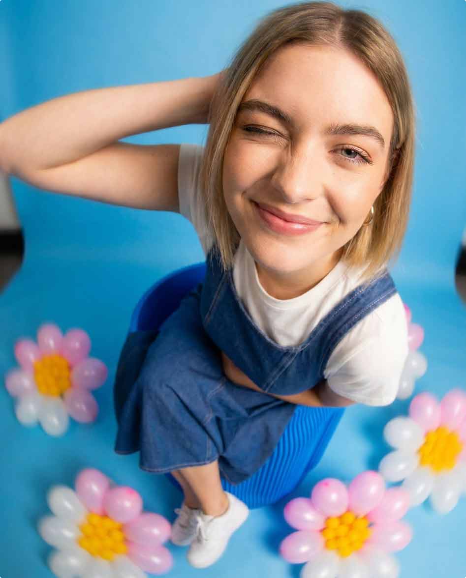 Junge Frau mit geschlossenem Auge lächelt, sitzend auf einem blauen Hocker umgeben von Blumenballons.