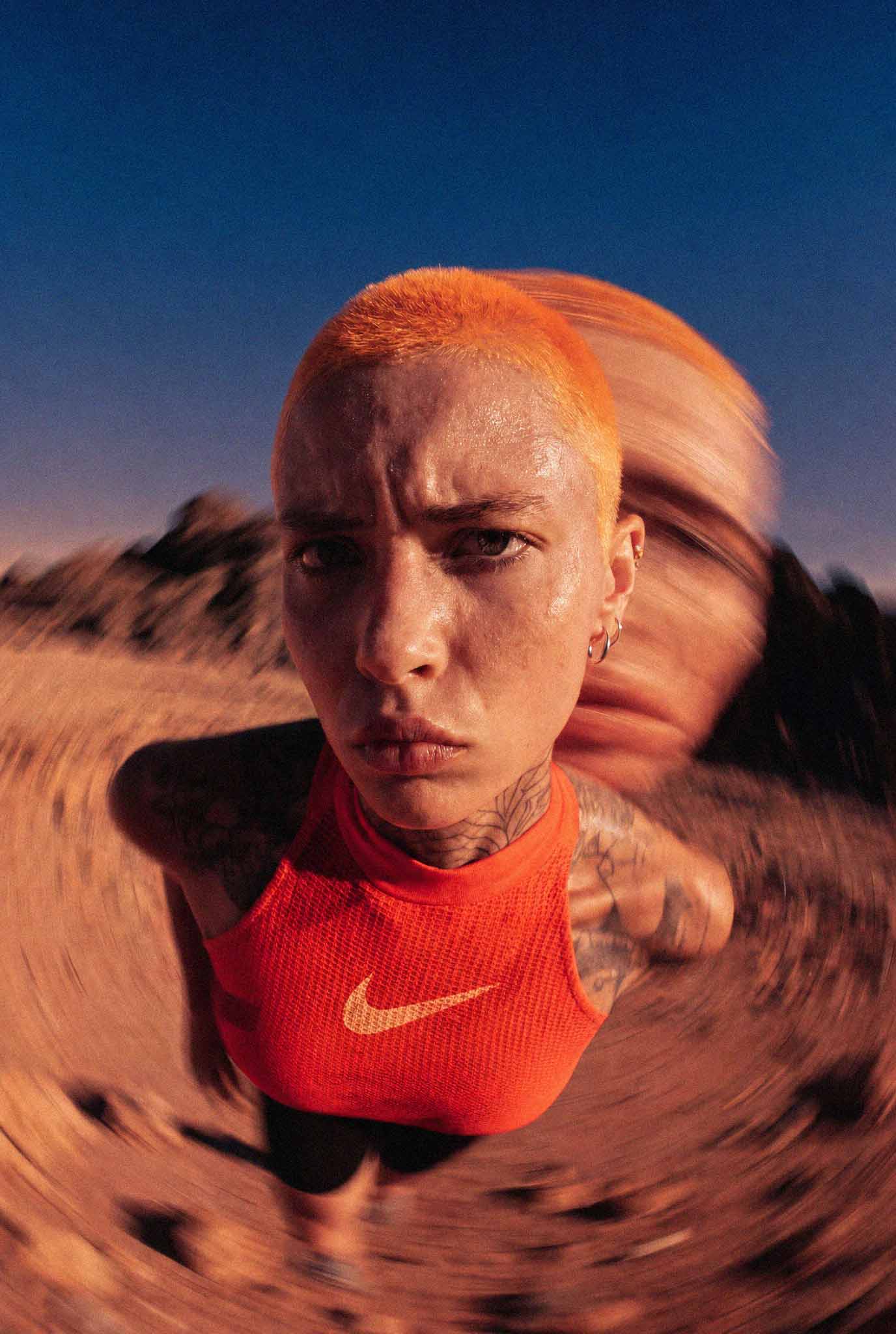 Nahaufnahme einer Person mit orangefarbenem Haarschnitt und rotem Nike-Oberteil in Wüstenumgebung.
